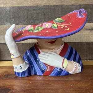 Peut inclure: Une figurine en céramique d'une femme portant un chapeau rouge et violet avec des détails floraux. La femme porte une robe à rayures bleues et blanches avec un gant blanc sur sa main gauche.