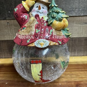 Könnte beinhalten: Eine Schneemann-Figur aus Keramik mit Cowboyhut und roter Fransenjacke, die einen grünen Tannenbaum hält. Der Schneemann sitzt auf einem klaren, kugelförmigen Glasgefäß mit Rissoptik, das mit einem bemalten gelb-roten Cowboystiefel verziert ist.