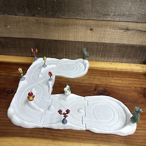Op de afbeelding: Een witte, met sneeuw bedekte miniatuur dorpsscène met een kronkelende weg, kleine bomen en decoratieve straatlantaarns. Een sneeuwpopfiguur en andere miniatuurfiguren zijn ook aanwezig. De scène staat op een houten oppervlak.