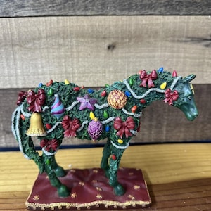 May include: A green horse ornament decorated for Christmas. The horse is adorned with red bows, colourful lights, and baubles. It stands on a red base with a gold trim. A festive holiday decoration.