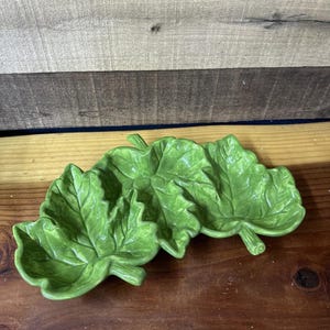 May include: A set of three green ceramic leaf-shaped serving dishes. The dishes are a bright, glossy green and have detailed veining to mimic real leaves. The dishes are arranged on a wooden surface.