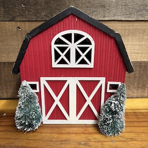 May include: A red wooden barn decoration with a black roof, white trim, and a white arched window. The structure features large white double doors with an X-pattern and is flanked by two small, snow-dusted green bottle brush pine trees.