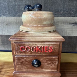May include: A brown ceramic cookie jar shaped like a vintage coffee grinder with a black handle on top. The jar has a drawer with a black knob and the word "COOKIES" written in red letters.