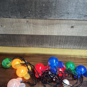 Puede incluir: Una cadena de luces multicolores con forma de cabeza de personaje de dibujos animados. Las luces son verdes, naranjas, rojas, azules y rosa iridiscente. Las luces están en un cable negro, sobre una superficie de madera.
