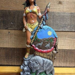 May include: A painted resin figurine featuring a person with long dark hair wearing a yellow top and skirt, standing beside a circular shield decorated with a landscape, a moon, and two wolves. A gray wolf stands on a rocky base below.