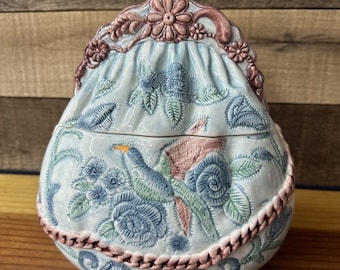 May include: A ceramic trinket box shaped like a purse with a floral design and a pink bird. The purse has a blue and white floral pattern with pink accents. The top of the purse is closed with a pink flower.