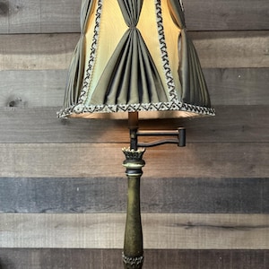 May include: An antique brass lamp with a pleated olive green and gold lampshade. The shade is trimmed with a decorative black and white border. The lamp has a detailed base and a pivoting arm.