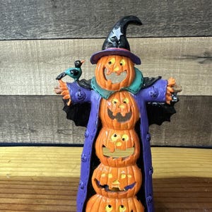May include: A Halloween-themed decorative figurine featuring a stack of carved orange pumpkins with smiling faces. The figurine is dressed in a purple coat and a witch's hat, with a small bird perched on its arm.