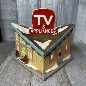 May include: A ceramic Christmas village house with a red and white sign that reads "TV & APPLIANCES". The house is decorated with snow and has a lit window.