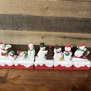 Könnte beinhalten: Eine Sammlung von Keramik-Schneemannfiguren, jede auf einem roten Sockel mit einer weißen, schneegleichen Oberseite. Die Schneemänner sind mit Hüten, Schals und anderen festlichen Details verziert.