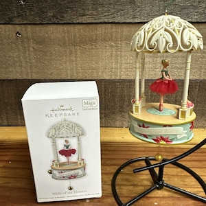May include: A Hallmark Keepsake Nutcracker Suite Waltz of the Flowers Christmas tree ornament featuring a ballerina in a red tutu inside a cream-colored gazebo structure, accompanied by its white product box with matching graphic imagery.