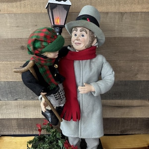 May include: A festive Christmas figurine set. One figure is in a grey coat, red scarf, and top hat, the other in a green and red plaid hat and scarf. A black and white lamppost stands between them. The base has festive greenery.