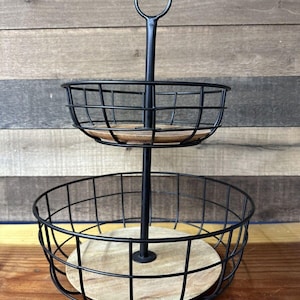 May include: A two-tiered black wire stand with wooden bases. The stand has a handle on the top and is perfect for displaying fruit or other items.