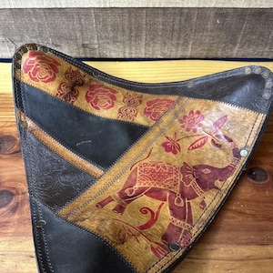 May include: Triangular leather stool with a decorative elephant and floral design. The stool features a patchwork design with black, tan, and brown leather. The elephant is red and tan, and the floral accents are red.