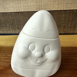 May include: A white ceramic candy corn shaped container with a smiling face. The container has a rounded top and a flat base. The face has large, round eyes, a small nose, and a curved smile. The container is unpainted.
