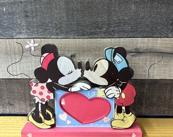 May include: Wooden Mickey and Minnie Mouse kissing photo frame. The frame features a pink base, a red heart-shaped photo holder, and the words "To" and "First Class Mail". Minnie wears a polka dot dress and Mickey wears a red shirt.