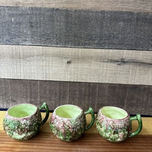 May include: Three small green and pink ceramic cups with floral designs and handles. The cups are sitting on a wooden surface.