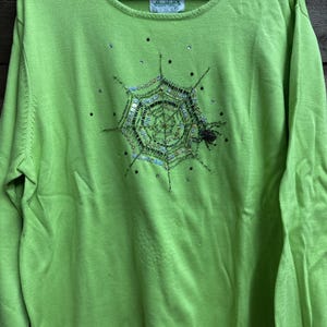 May include: A lime green long-sleeve sweater featuring a central spider web design embellished with green and silver sequins, beads, and a small black spider. The garment has a round neckline and a small white brand label at the inner collar.