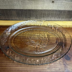 May include: An oval clear glass serving platter featuring a molded turkey design in the center. The rim of the platter is decorated with a raised relief pattern of various fruits, including grapes and pears, and small floral accents.