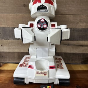 Pode incluir: Robô de brinquedo branco e vermelho com uma base tipo tanque. O robô tem uma viseira vermelha e um rosto preto com quatro botões. O robô é etiquetado como "RAD" na base. Inclui duas baterias pretas.