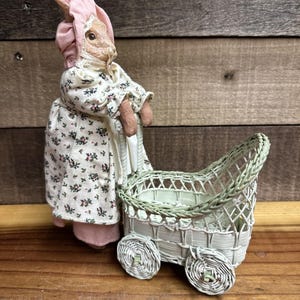 May include: A vintage bunny figurine wearing a pink bonnet and floral dress, pushing a light green wicker stroller with woven wheels. The bunny has a detailed face and is standing on a wooden surface.