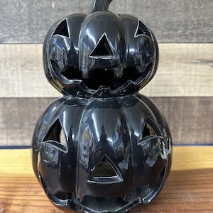 Könnte beinhalten: Ein schwarzer Kerzenhalter aus Keramik in Form eines Kürbisses mit einem geschnitzten Jack-o'-Lantern-Gesicht. Der Kürbis ist auf einem anderen Kürbis mit ähnlichem Design gestapelt.