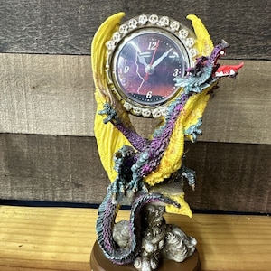 May include: A purple and yellow dragon figurine with a clock face on its back. The clock face has a black background with a red lightning bolt and white numbers. The dragon is perched on a rock base with a brown base.