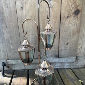 Peut inclure: Une lampe d'extérieur à trois lanternes de couleur bronze. Les lanternes sont hexagonales avec des panneaux de verre et sont suspendues à des bras métalliques incurvés. La lampe a une base circulaire et est placée sur un fond en bois.