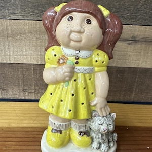 Peut inclure: Figurine en céramique d'une fille en robe jaune à pois noirs, tenant une fleur et caressant un chat gris. La fille a les cheveux bruns en couettes avec des nœuds jaunes. La figurine est sur une base blanche.