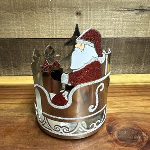 May include: A Christmas-themed candle holder featuring Santa Claus in a sleigh. The holder is metallic with silver glitter accents. Santa is dressed in a red suit and hat with white trim. A Christmas tree and gifts are also visible.