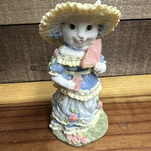 May include: A white rabbit figurine wearing a yellow hat and a blue dress with pink and blue floral accents. The rabbit holds a pink fan and a small card. The figurine is standing on a patch of green grass.