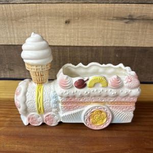 May include: A whimsical, white ceramic planter shaped like a train, featuring an ice cream cone chimney. The train car is decorated with pink, yellow, and blue accents, and topped with frosting and fruit details. The wheels have yellow flower centers.
