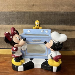May include: A ceramic Mickey and Minnie Mouse figurine set with a light blue oven in the center. Minnie wears a red polka dot bow and dress. Mickey wears a chef's hat. Pluto sits on top of the oven.