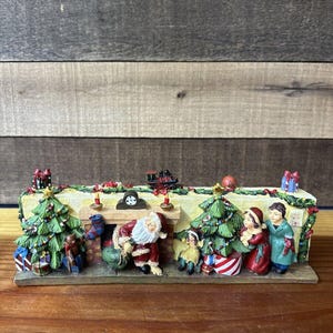 Peut inclure: Une figurine de scène de Noël avec le Père Noël, des enfants et une cheminée. La figurine est en résine peinte et présente un design détaillé avec une horloge, un train et un sapin de Noël.