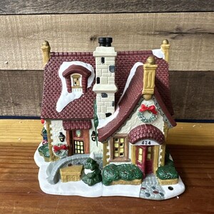 May include: A ceramic Christmas village house with a red roof, white snow, and a wreath on the door. The house has a number "424" on the door.