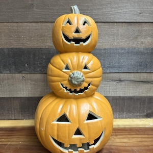 Può includere: Decorazione a forma di zucca impilata con tre facce di jack-o'-lantern intagliate. Le zucche sono arancioni con occhi triangolari neri e sorrisi sdentati. La zucca in alto ha un gambo verde.
