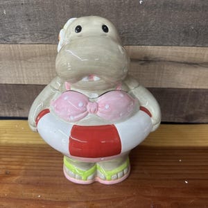 Könnte beinhalten: Eine Keramik-Nilpferdfigur mit einem rosa Bikini mit Punkten und einer weiß-roten Schwimmweste. Das Nilpferd hat eine Blume im Haar und grüne und rosa Flip-Flops. Die Figur steht auf einer Holzoberfläche.