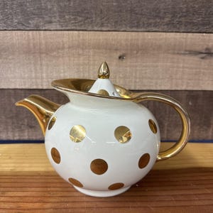 May include: A white ceramic teapot with gold polka dots and gold trim. The teapot has a gold handle, spout, and lid. The lid is topped with a gold finial. The teapot is a classic design.
