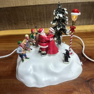 Peut inclure: Une scène de village de Noël avec le Père Noël et une femme dansant, un groupe de musique, des pingouins et un lampadaire. Les figurines sont sur une base enneigée avec un arbre enneigé.