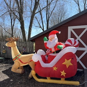 Könnte beinhalten: Eine aufblasbare Weihnachtsdekoration mit dem Weihnachtsmann in einem roten Schlitten, der von einem Rentier gezogen wird. Der Schlitten hat goldene Sterne und weiße Akzente. Das Rentier ist hellbraun mit einem grünen und roten Geschirr.