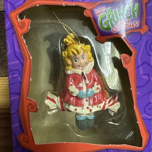 May include: A Cindy Lou Who Christmas ornament from Dr. Seuss How The Grinch Stole Christmas, featuring a blonde character in a red coat with white polka dots and blue mittens, packaged in a purple and orange box with a gold hanging string.