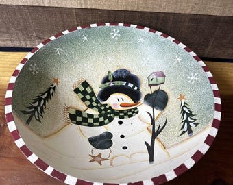 May include: A wooden bowl with a hand-painted snowman design. The snowman is wearing a black hat, green scarf, and has a red nose. The bowl has a red and white striped rim.