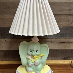 May include: A whimsical table lamp featuring a light green elephant figurine base with a yellow bow and a pink hat. The lamp has a cream-colored pleated lampshade. The base is on a yellow and pink circular platform.