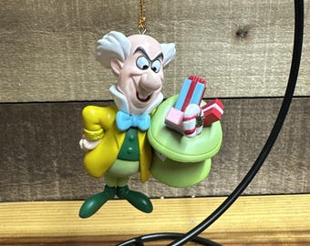 May include: A colorful ornament featuring a cartoon character in a yellow jacket, green vest, and green shoes. The character holds a green hat with small gift boxes. The ornament has a gold string for hanging and is set against a wooden background.
