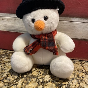 Könnte beinhalten: Weißer Plüsch-Schneemann mit einem schwarzen Hut und einem rot-schwarz karierten Schal. Der Schneemann hat eine Karottennase und schwarze Augen.