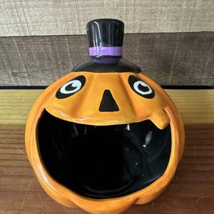 Könnte beinhalten: Orangefarbener Keramikkürbis mit einem schwarzen Zylinder und einem violetten Band. Der Kürbis hat einen schwarzen Mund und weiße Augen. Es ist eine Halloween-spezifische Süßigkeitenschale.