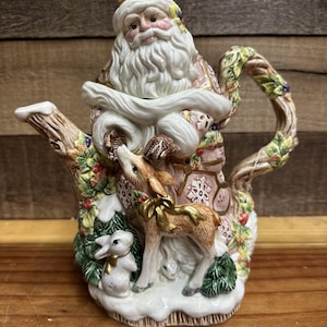 May include: A ceramic Christmas teapot depicting Santa Claus, a deer, and a rabbit. The teapot is decorated with pink, white, and gold details. The handle is designed to resemble a tree branch, and the spout is shaped like a tree trunk.