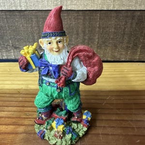 Puede incluir: Figura de gnomo navideño con gorro rojo, pantalón verde y barba blanca. El gnomo sostiene una caja de regalo azul con un lazo amarillo y un saco rojo. La base está decorada con regalos y vegetación.