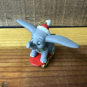 May include: A grey Dumbo figurine wearing a Santa hat and a green and red scarf, riding a red sled with gold trim. The figurine has large ears and blue eyes. A festive Christmas ornament.