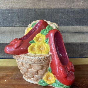 Puede incluir: Zapatos rojos de cerámica con flores amarillas y hojas verdes en una cesta de mimbre marrón.